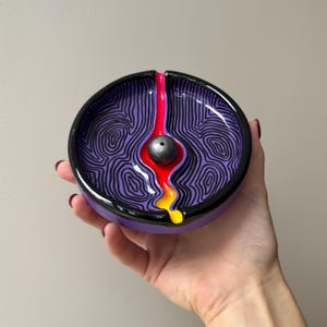 Puede incluir: Un porta-incienso redondo de cerámica con interior morado y patrones de líneas topográficas negras. Una esfera metálica central se sitúa en un canal rojo y amarillo que fluye hacia el borde negro del plato circular.
