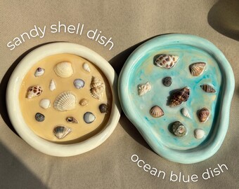 Puede incluir: Dos platos redondos con bordes elevados, uno beige etiquetado como sandy shell dish y otro azul claro etiquetado como ocean blue dish. Ambos platos están rellenos con una variedad de pequeñas conchas naturales incrustadas en un acabado liso y brillante.