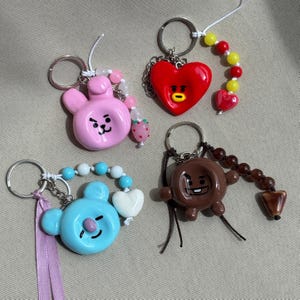 Op de afbeelding: Vier sleutelhangers met cartoonfiguur-bedels: een roze konijn, een rood hart, een lichtblauwe koala en een bruin wezen. Elke hanger heeft een metalen ring, een snoer met kleurrijke kralen en extra accenten zoals een aardbei of een lint.