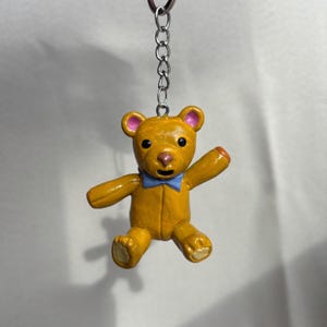 May include: A small, yellow teddy bear keychain with a blue bow tie. The bear has pink accents on its ears and nose, and orange accents on its paws. The keychain is attached to a silver chain.
