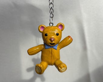 May include: A small, yellow teddy bear keyring with a blue bow tie. The bear has pink accents on its ears and nose, and orange accents on its paws. The keyring is attached to a silver chain.