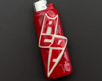 May include: A red lighter with the word "ACE" in white, stylized lettering. The lighter has a glossy finish and a silver metal top. The design is centered on the lighter's body.