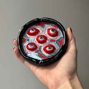 May include: A black ashtray with a glossy finish, featuring a unique design of multiple red, white, and blue eyeballs. The ashtray is round with a recessed area for holding cigarettes. The design is artistic and eye-catching.