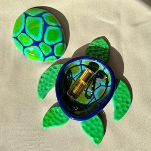 May include: A handcrafted, decorative turtle-shaped trinket dish with a matching lid. The dish is primarily green and blue, with a gold-colored bottle and a black beaded necklace inside. The lid is a painted stone with a similar design.