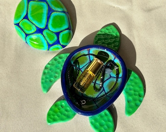 May include: A decorative turtle-shaped trinket dish with a matching lid. The dish is green and blue, with a gold-coloured bottle and a black beaded necklace inside. The lid is a painted stone with a similar design. The dish is likely made of ceramic.