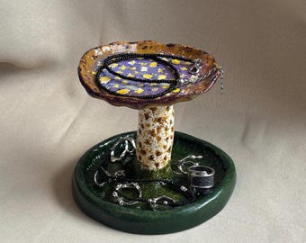 Puede incluir: Un soporte para joyas de cerámica con forma de seta, base verde oscuro, tallo blanco moteado y sombrero morado con motivos florales amarillos. La parte superior sostiene un collar de cuentas negras y la base contiene varios anillos plateados.