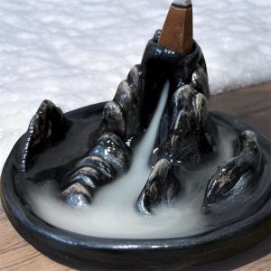 Pode incluir: Um porta-incenso de cerâmica preta em forma de cordilheira. O porta-incenso tem um acabamento escuro e brilhante com um efeito de cachoeira de fumaça branca. Um cone de incenso marrom está no topo da montanha.