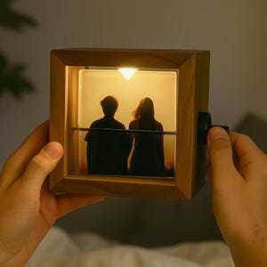 Puede incluir: Un marco de fotos de madera con una luz cálida que ilumina una foto de dos personas. El marco tiene forma cuadrada con un pequeño botón negro en el lateral. La imagen interior muestra las siluetas de dos personas. El marco está sostenido por las manos de una persona.