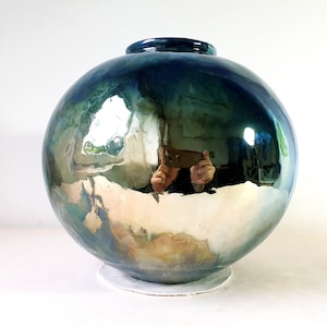 Op de afbeelding: Een grote, ronde, blauwe en groene keramische vaas met een metallic afwerking. De vaas heeft een reflecterend oppervlak dat een vervormde reflectie van de omgeving laat zien.