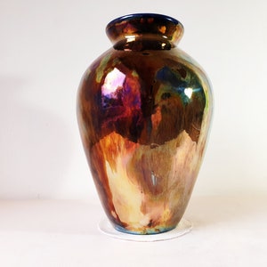 Puede incluir: Un jarrón grande iridiscente con un esmalte marrón, dorado y azul. El jarrón tiene un cuerpo redondo y un cuello estrecho.
