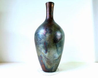 May include: A tall, slender ceramic vase with a rounded base and a narrow neck. The vase has a metallic glaze with shades of blue, green, and copper.
