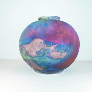 Puede incluir: Un jarrón redondo de cerámica con un esmalte iridiscente vibrante en tonos de azul, verde, rosa y morado. El jarrón tiene un acabado liso y brillante.