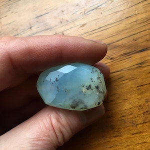May include: A faceted, oval-shaped gemstone with a pale blue hue and dark inclusions. The stone is held in a hand, showcasing its polished surface and unique internal patterns. The gemstone is a natural, organic material.