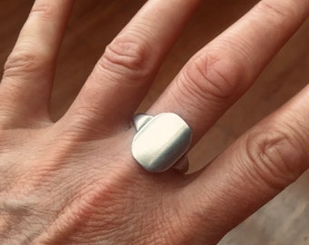 May include: A silver ring with a rectangular face. The ring is worn on a finger, showcasing its simple, modern design. The metal has a brushed finish, reflecting light. The ring's band is thin, complementing the larger face.