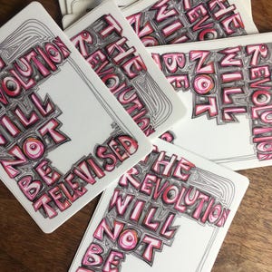 May include: A stack of white cards with the text "THE REVOLUTION WILL NOT BE TELEVISED" in pink and black lettering. The cards are arranged on a wooden surface.