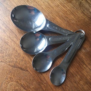 May include: Set of four stainless steel measuring spoons on a wooden surface. The spoons are attached with a metal ring and have measurements of 15 ml, 5 ml, 2.5 ml, and 1.25 ml. The spoons are angled towards the viewer.