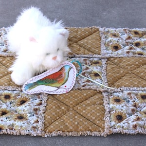 May include: A white plush cat toy rests on a patchwork pet bed. The bed features a sunflower and brown floral pattern. A colorful bird-shaped tag is attached to the bed with a white string. The bed is approximately 18 inches by 14 inches.
