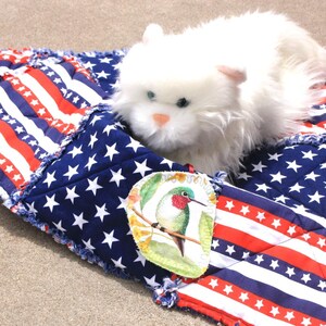 May include: A white plush cat rests on a patriotic, quilted pet bed. The bed features a design of red and white stripes and blue fields with white stars, resembling the American flag. A small, colorful hummingbird patch is sewn on the bed.