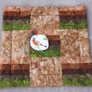 May include: A handmade, square quilted placemat with a patchwork design in shades of brown, green, and tan. A decorative tag with a bird illustration is attached with a button and green ribbon. The placemat is likely used for dining or home decor.