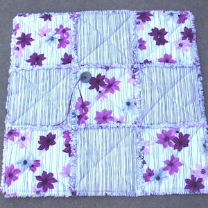 May include: Square quilted patchwork blanket with a floral pattern in shades of purple and white. The blanket features a button closure and a striped pattern in between the floral squares. The edges have a frayed, textured finish.