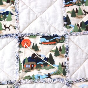 May include: A handmade quilt featuring a camping theme. The quilt is composed of square patches with a white background and camping scenes with tents, trees, and mountains. The edges are trimmed with a blue and white frayed fabric. An orange button secures the quilt.