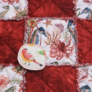 May include: A red and white patchwork quilt with a variety of sea-themed fabric squares. One square features a colorful bird with a red beak and yellow body. The quilt is decorated with a button.