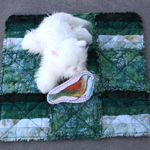 May include: A white plush cat rests on a green and white quilted mat. A decorative bird-shaped tag with a colorful illustration is placed on the mat. The mat has a patchwork design with various shades of green.