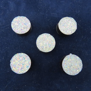 May include: Five round, white druzy plugs with iridescent sparkle are arranged on a dark blue surface. The plugs have a flat, black backing. The druzy surface is textured and reflects light, creating a shimmering effect.