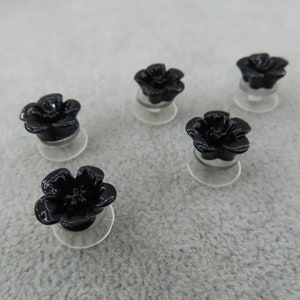 May include: Five black flower-shaped ear plugs. Each plug has a glossy finish and sits on a clear, circular base. The plugs are arranged on a light gray surface, showcasing their floral design.