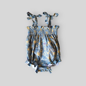 May include: A blue romper featuring a yellow and white floral pattern, a smocked bodice with a ruffled top edge, and two shoulder straps that tie into bows. The leg openings are finished with a ruffled trim, creating a gathered silhouette.
