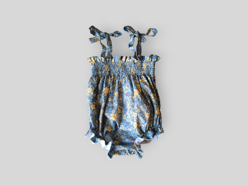 May include: A blue romper featuring a yellow and white floral pattern, a smocked bodice with a ruffled top edge, and two shoulder straps that tie into bows. The leg openings are finished with a ruffled trim, creating a gathered silhouette.