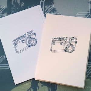 May include: Two blank greeting cards featuring a blue line drawing of a vintage camera. The cards are white and off-white, with the camera design centred on the front. The background has a blue and white geometric pattern.
