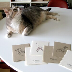 May include: A grey tabby cat rests on a white table, gazing upwards. Several square greeting cards with chair designs are arranged on the table. The cards feature line drawings of iconic chair designs in black and purple.