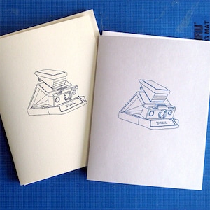 Peut inclure: Deux cartes de vœux vierges avec un dessin au trait bleu d'un appareil photo instantané sur chaque carte. Les cartes sont sur un fond de grille bleue.