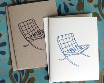 May include: Greeting cards featuring a minimalist line drawing of a modern chair design. One card is printed in black ink on a tan card, and another is printed in blue ink on a white card. The cards are stacked on a patterned blue and gold surface.