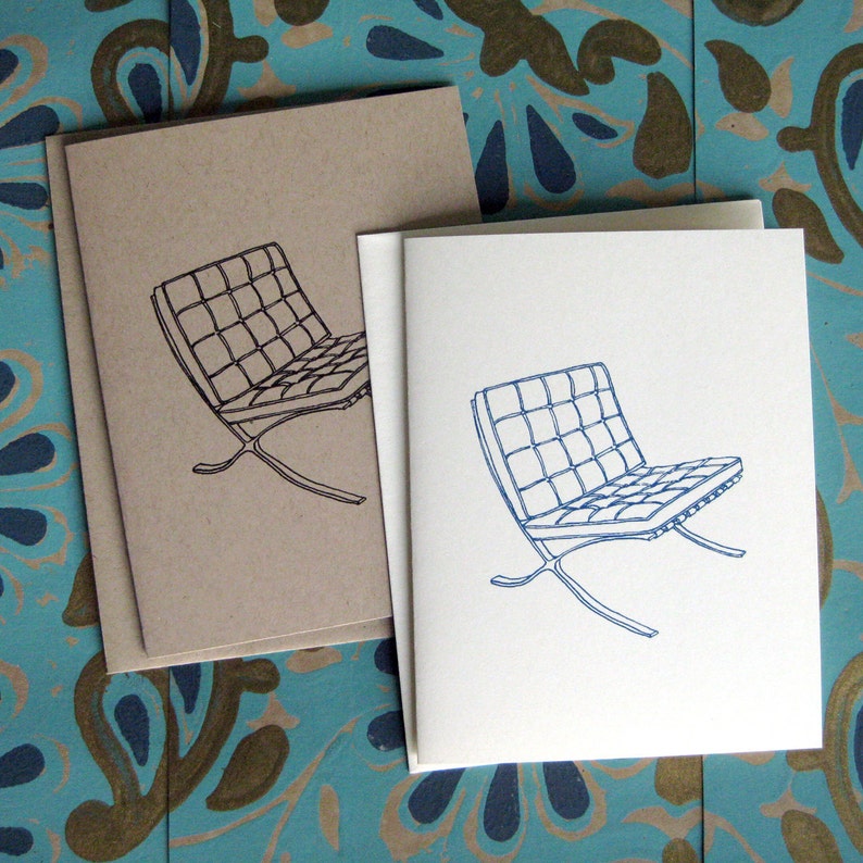 May include: Greeting cards with a minimalist line drawing of a modern chair design. One card is printed in black ink on a tan card, and another is printed in blue ink on a white card. The cards are stacked on a patterned blue and gold surface.