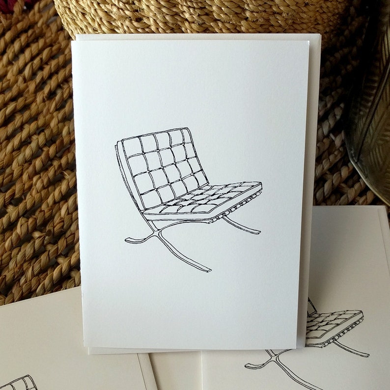 May include: A pile of white greeting cards, each with a black line drawing of a modern chair. The chair design includes a rectangular, grid-patterned seat and a curved metal base. The cards are set against a neutral backdrop.