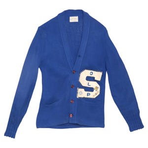 Könnte beinhalten: Eine königsblaue Strickjacke mit Schalkragen, langen Ärmeln mit gerippten Bündchen und vier braunen Knöpfen. Ein großer weißer Aufnäher auf der Vorderseite zeigt den Buchstaben S mit den Buchstaben O, L und P sowie eine kleine Fußballgrafik.