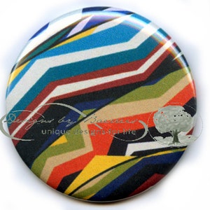 May include: A round button with a colorful geometric design. The pattern features zig-zag lines in shades of blue, teal, red, orange, yellow, green, and black. The button has the text "Designs by Baerssel" and "unique designs for life".
