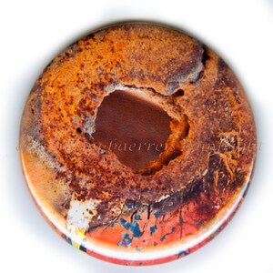 May include: A round, weathered button with a large central hole. The button features a textured surface with shades of orange, brown, and red, suggesting rust or aged metal. The edges show hints of blue and white.