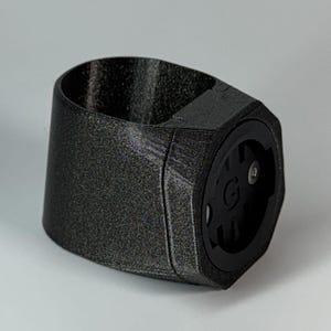 Può includere: Un supporto per luci da bicicletta stampato in 3D nero con una superficie testurizzata. Il supporto ha una forma esagonale e un'apertura circolare per una luce. Il supporto ha una piccola vite sul lato.