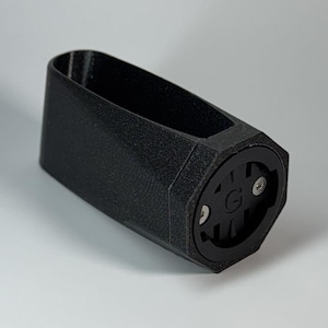 Könnte beinhalten: Ein schwarzer, 3D-gedruckter Fahrradcomputerhalter. Die Halterung hat einen rechteckigen Körper mit einer sechseckigen Basis, die ein kreisförmiges Design mit dem Buchstaben "G" in der Mitte und zwei silbernen Schrauben aufweist.