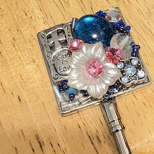 May include: A decorative silver key featuring a square top adorned with a blue gem, pink and clear crystals, and a white flower with a pink center. The text "God is Love" is visible on the top.