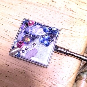 May include: A square, silver-toned purse hook with a decorative top. The top is embellished with purple, pink, and blue gems and beads. The hook is resting on a wooden surface.