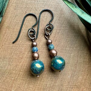 May include: A pair of dangle earrings with a teal and copper color scheme. The earrings feature a large teal bead with a copper bead and a smaller teal bead above it. The earrings are attached to a silver hook.
