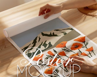 May include: A poster mockup featuring a mountain landscape with a field of orange poppies. The poster is rolled up and being held by a hand.