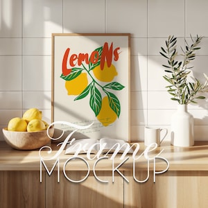以下が含まれることがあります： 白い背景に、緑の葉と「Lemons」という赤い文字とオレンジの文字で書かれたレモンのスタイリッシュなイラストが描かれた額装されたプリント。プリントは、レモンの入ったボウル、白いマグカップ、本が置かれた木製のカウンタートップの上に置かれています。
