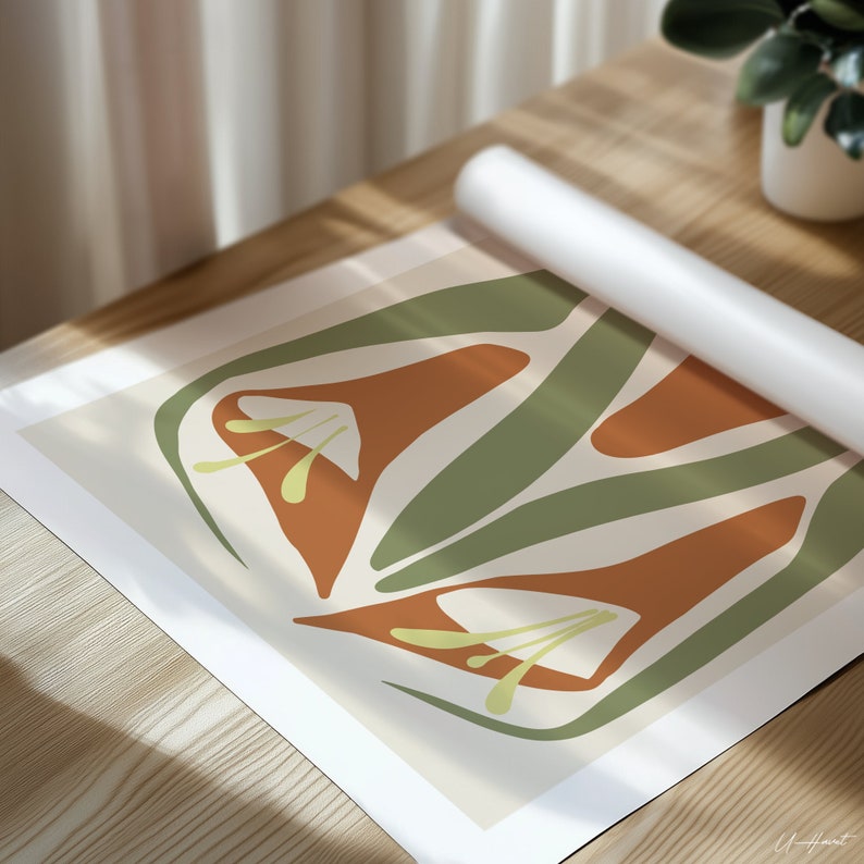 May include: An abstract art print featuring stylized orange and green floral shapes on a cream background. The print is partially rolled up, revealing the design. The artwork is displayed on a wooden surface, with a white roll of paper and a potted plant in the background.