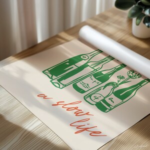 May include: A rolled-up white poster rests on a wooden surface, partially obscuring a beige print. The print features green line drawings of wine bottles and the orange text "a slow life."