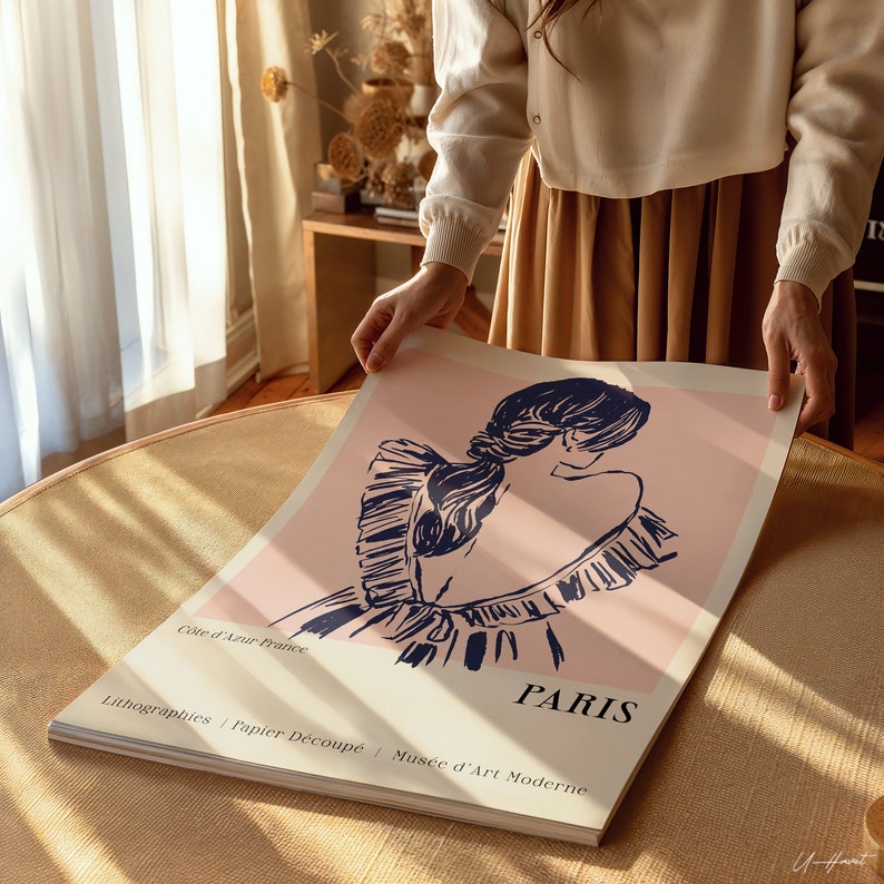Op de afbeelding: Een roze en blauwe poster met een gestileerde illustratie van een vrouw met haar haar naar achteren getrokken. De poster heeft de tekst "C&ocirc;te d'Azur France", "PARIS", "Lithographies | Papier D&eacute;coup&eacute; | Mus&eacute;e d'Art Moderne" en "12".